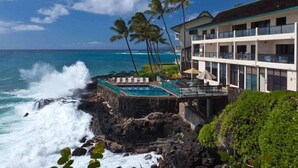 A hotel with a pool overlooking the ocean, palm trees, and a rocky shoreline.