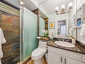 A modern bathroom with a glass shower enclosure, a white toilet, a sink with a marble countertop, and a large mirror above the vanity. - 3 Bedroom Deluxe Oceanfront with A/C - Poipu Shores 302B (Koloa)