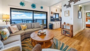 A spacious living area with a large window overlooking the ocean, a ceiling fan, and a cozy seating arrangement.