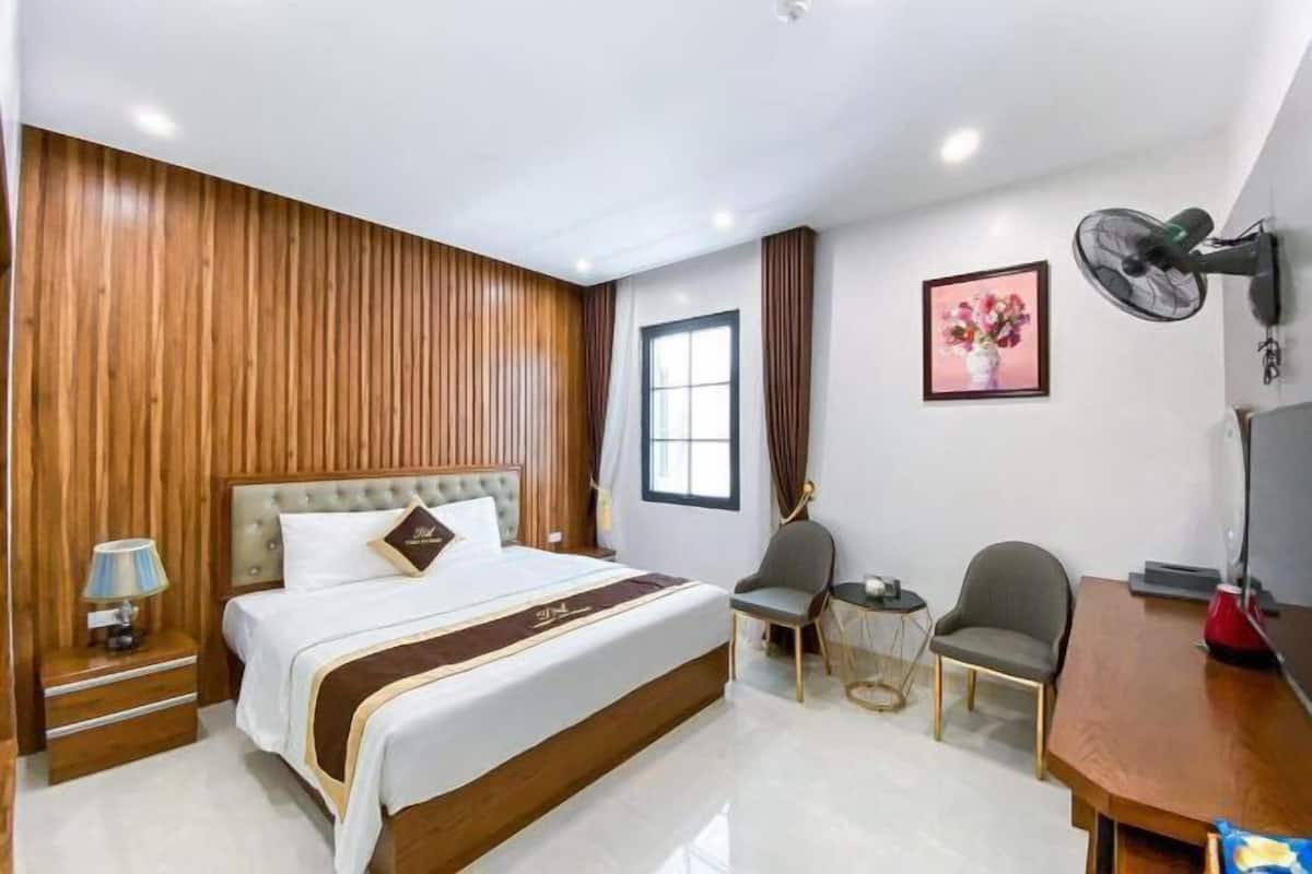 A hotel room with a bed, bedside table, lamp, chair, desk, fan, and a picture on the wall.