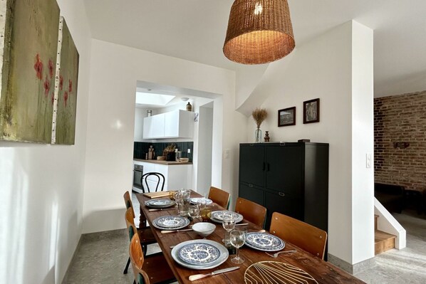 A dining area with a wooden table set for four, surrounded by chairs. There is a black cabinet to the right and a hanging light fixture above.