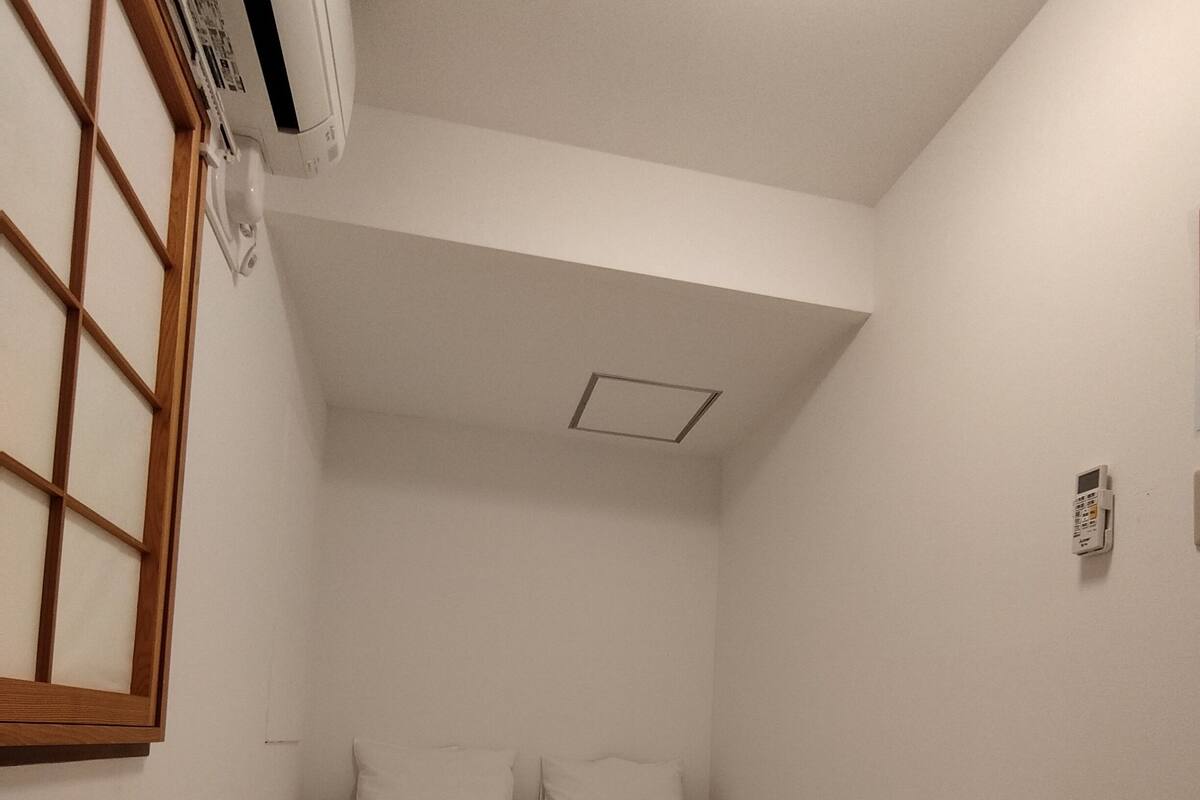 A bedroom with a bed, wooden flooring, a window with a grid pattern, and a ceiling-mounted air conditioner.