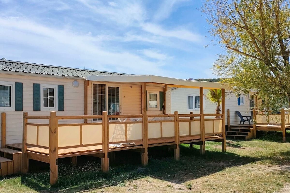 A mobile home with a wooden deck, a covered patio, and a tree in the background.