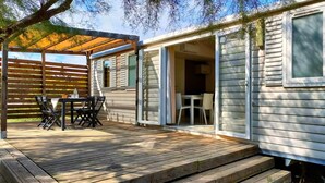 A wooden deck with a table and chairs, a covered patio area, and a sliding glass door leading to an interior space. - Camping Felix de la Bastide (Saint-Mitre-les-Remparts)
