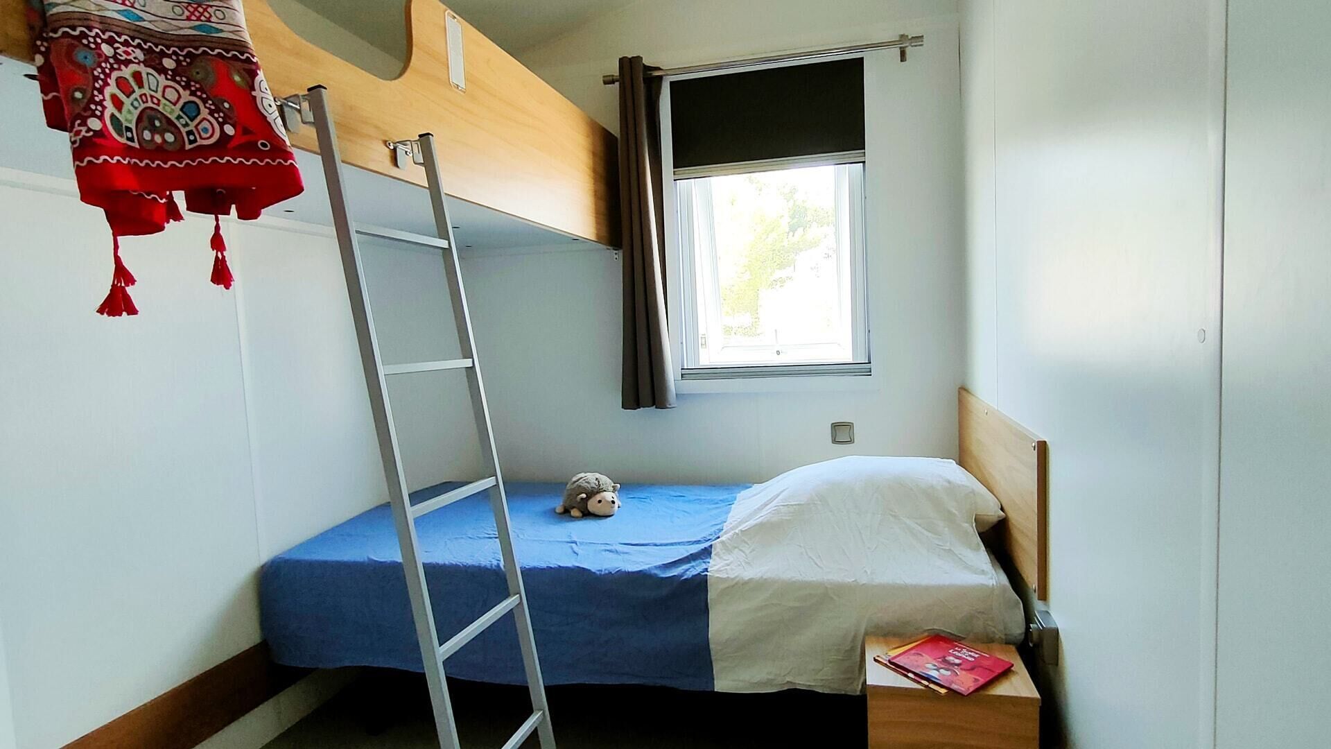 A small, compact room with a bunk bed, a single bed, a window with curtains, a ladder, and a small stuffed toy on the bunk bed.