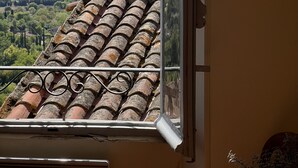 Vista a través de una ventana de un edificio con techo de tejas y chimenea, sobre un paisaje verde con colinas.
