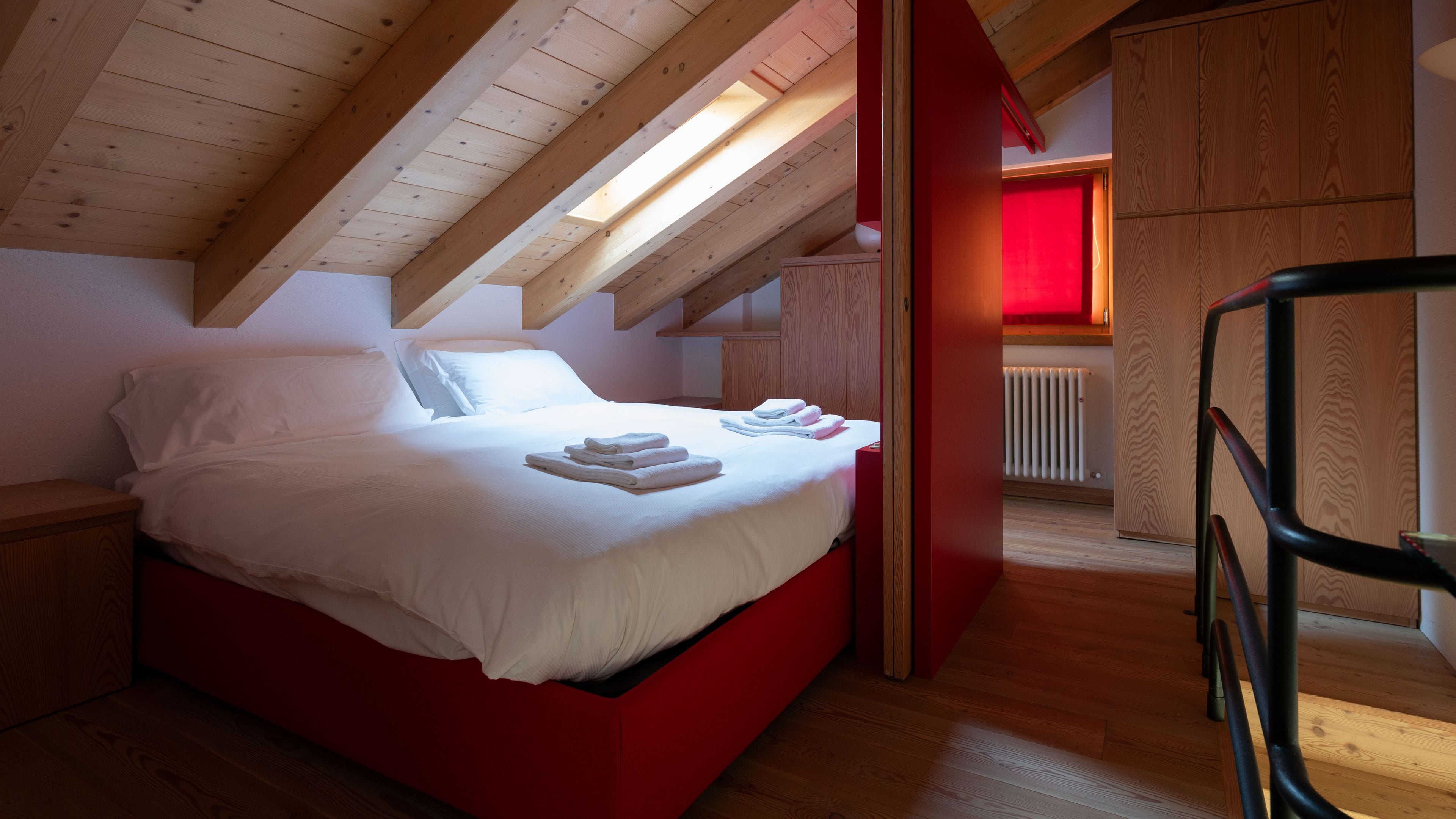 A bedroom with a red bed, white linens, a wooden floor, a skylight, and a red wall.