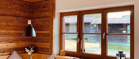 Un interior de cabaña acogedor con techo de madera, una mesa de comedor con sillas, un ventanal con vistas al exterior y un radiador debajo de la ventana.