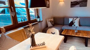 A cozy living room with a wooden ceiling, a sofa with decorative pillows, a dining table with a vase and a sign, and a view of a parked car through large windows. - Neu Renovierte Ferienwohnung für Bergverliebte (Reichenbach Oberstdorf)