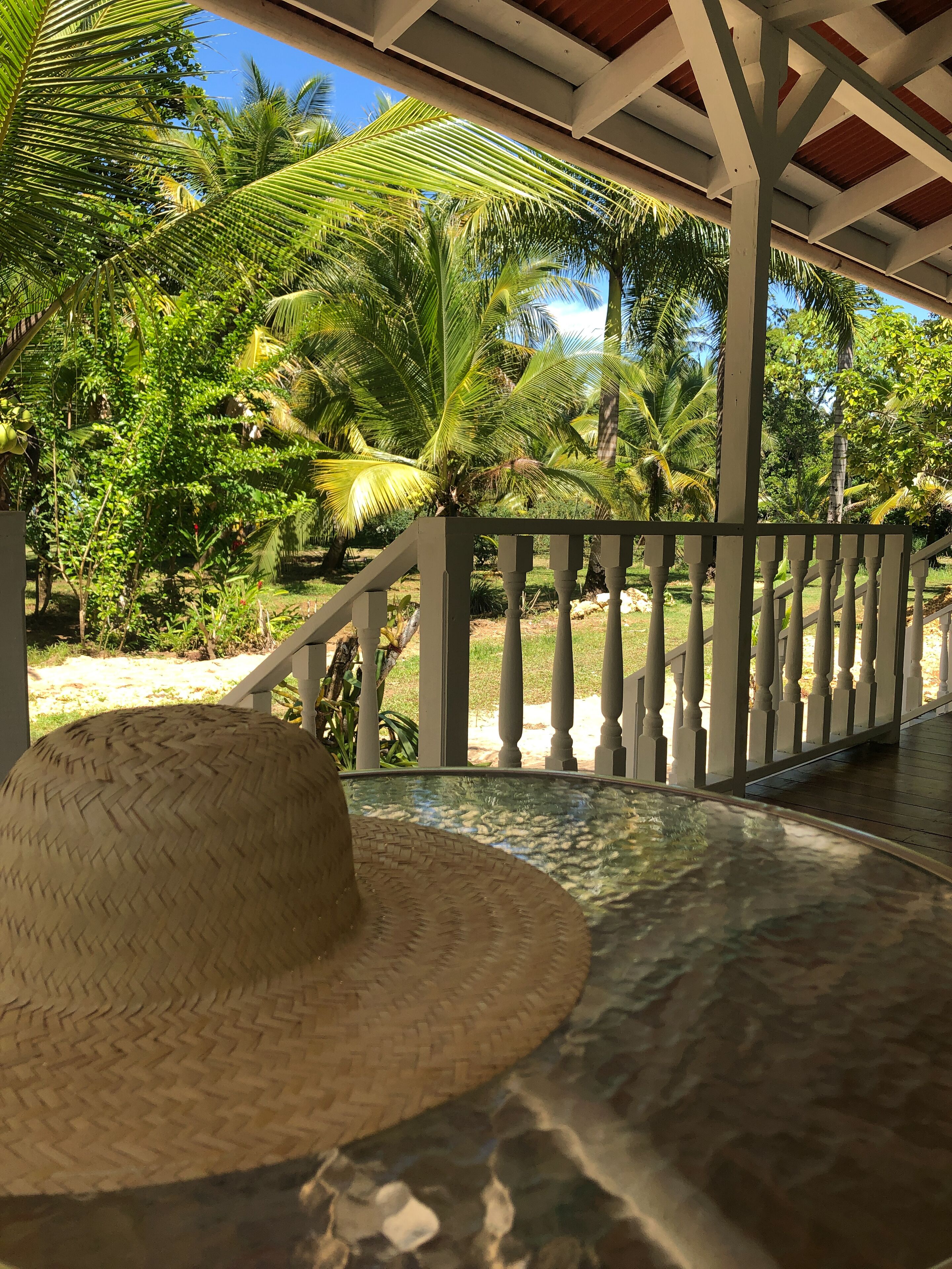 A covered outdoor area with a white railing, a woven hat on the floor, and lush greenery in the background.