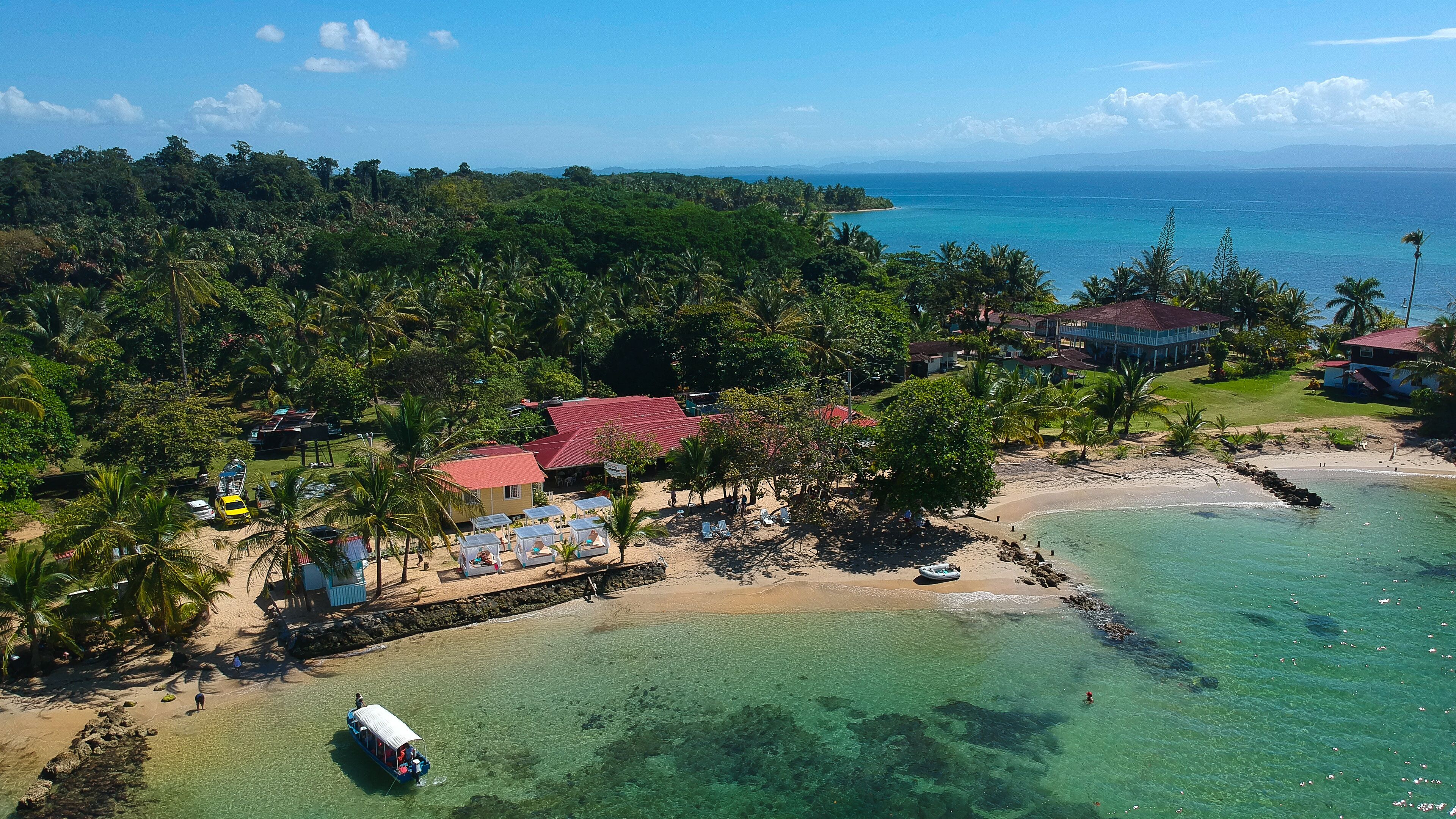 An aerial view of a tropical beach resort with clear waters, palm trees, and a small boat docked near the shore.