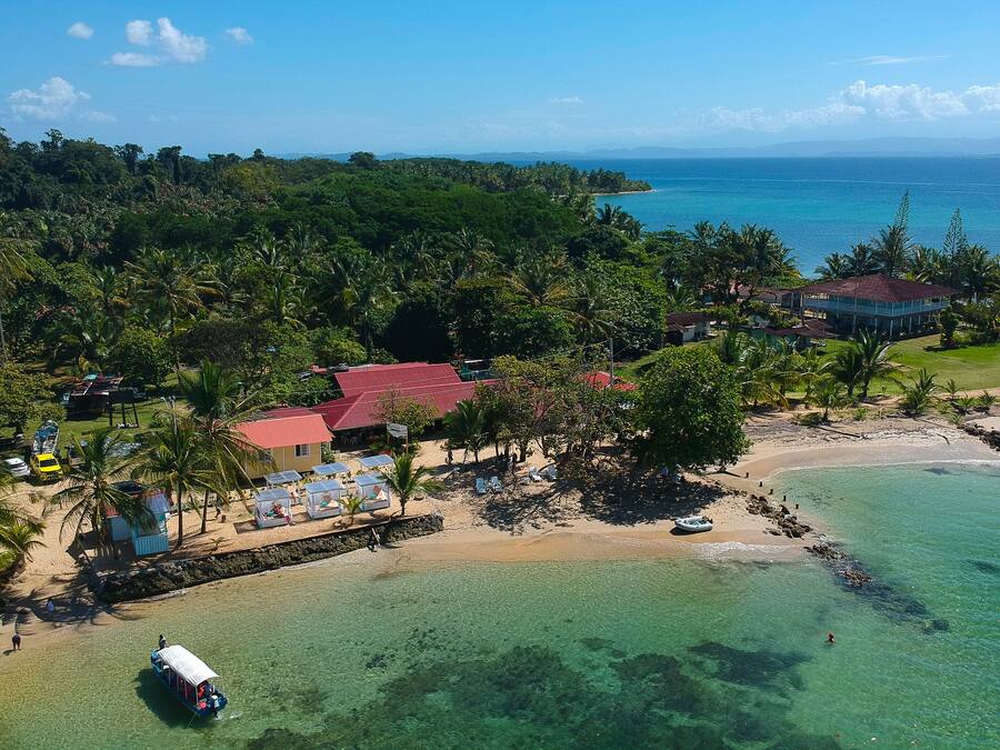 An aerial view of a tropical beach resort with clear waters, palm trees, and a small boat docked near the shore.