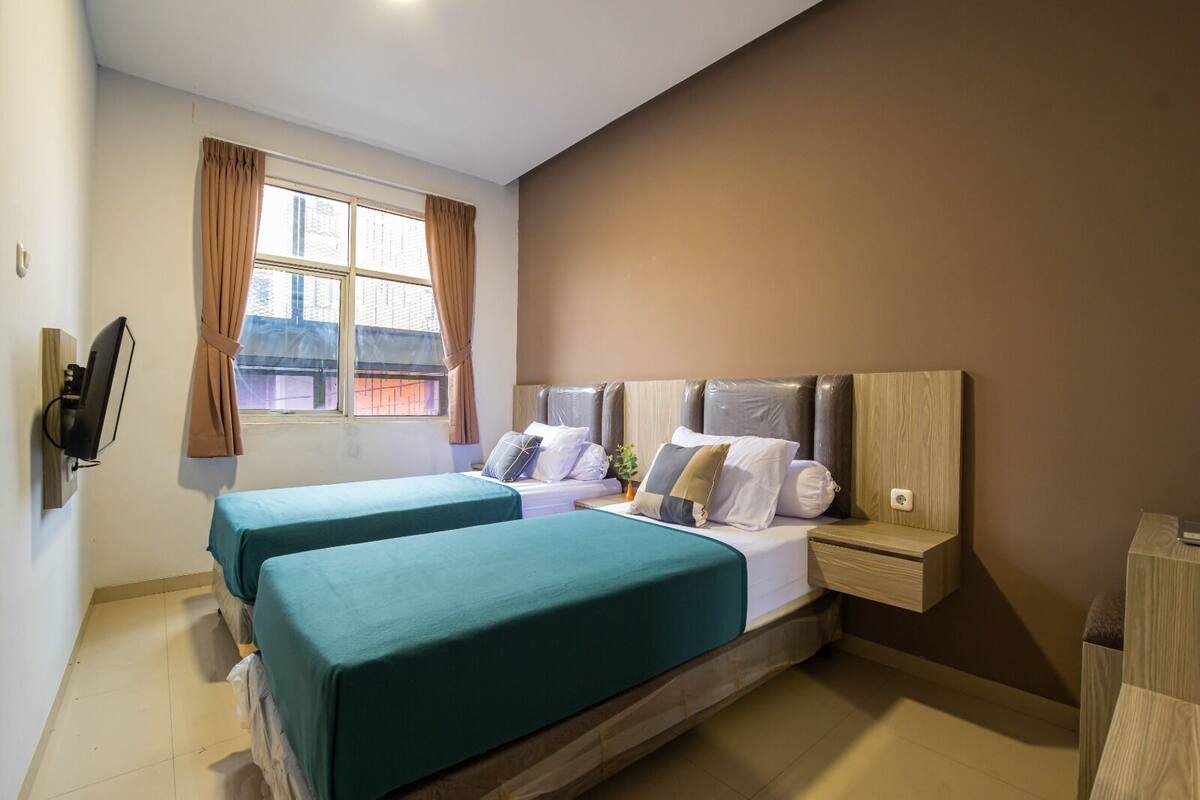 A hotel room with two beds, a TV mounted on the wall, a window with curtains, and a bedside table with a lamp.