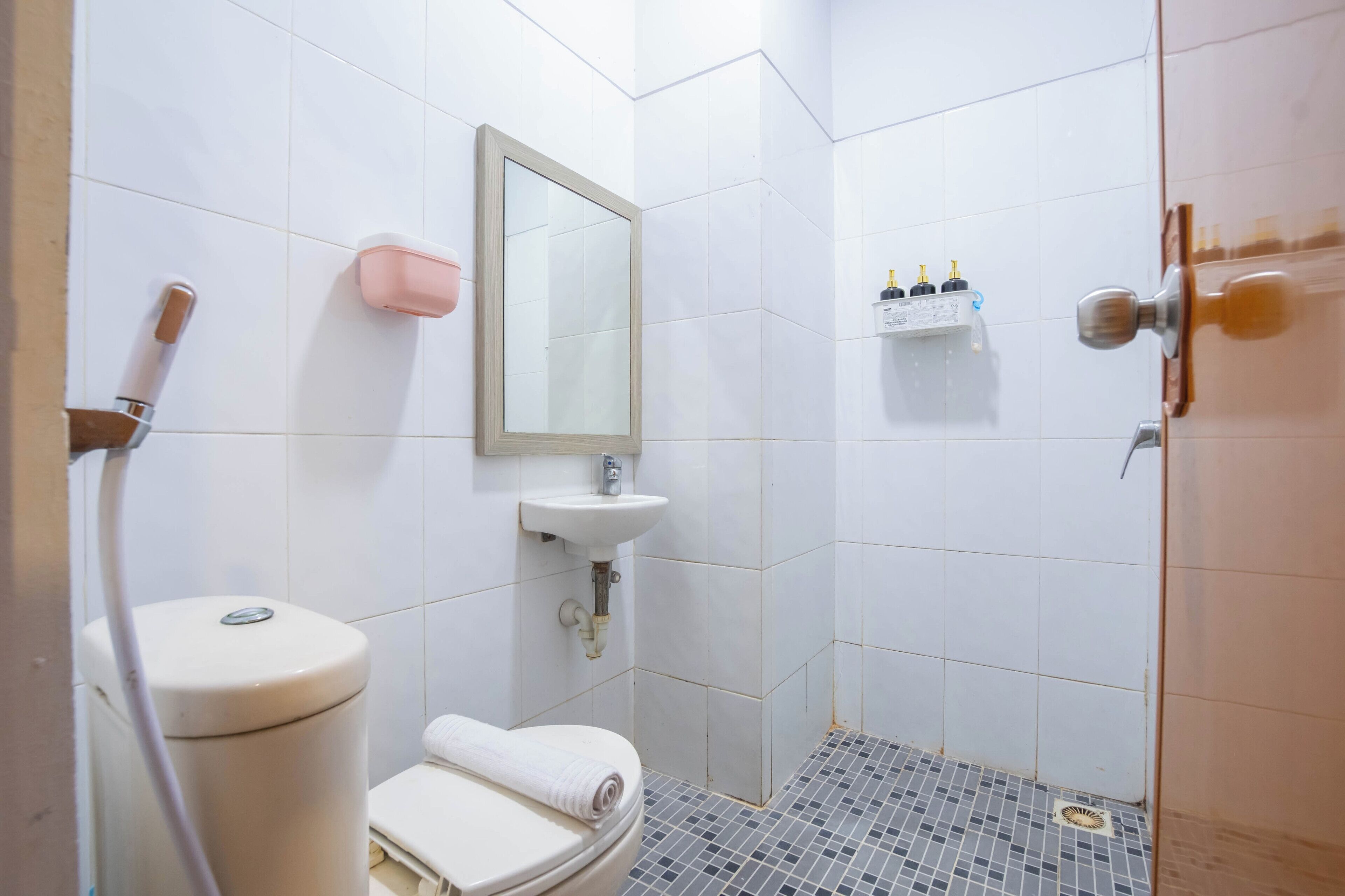 A bathroom with a toilet, sink, mirror, shower, and a small shelf with toiletries.