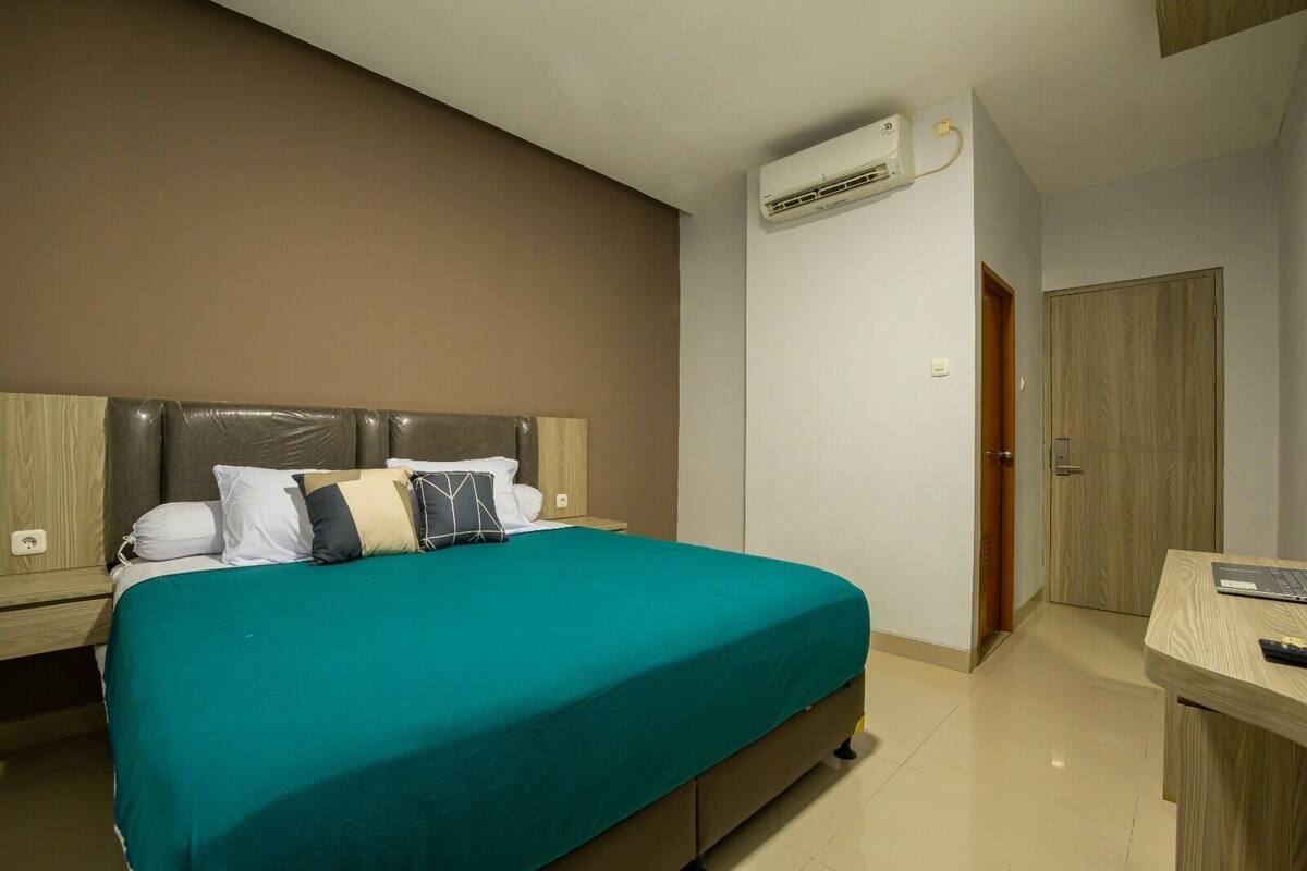 A hotel room with a bed, a desk, an air conditioning unit, and a wooden door.
