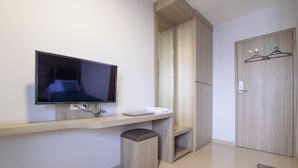 A modern hotel room with a flat-screen TV mounted on the wall, a wooden desk with a laptop, a small ottoman, and a wardrobe with a coat rack.