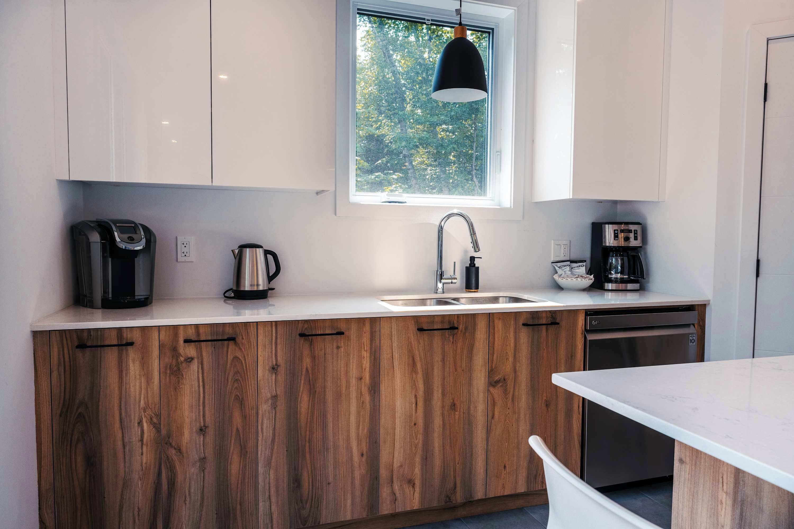 A modern kitchen with wooden cabinets, a stainless steel sink, a coffee maker, and a window with a view of trees.