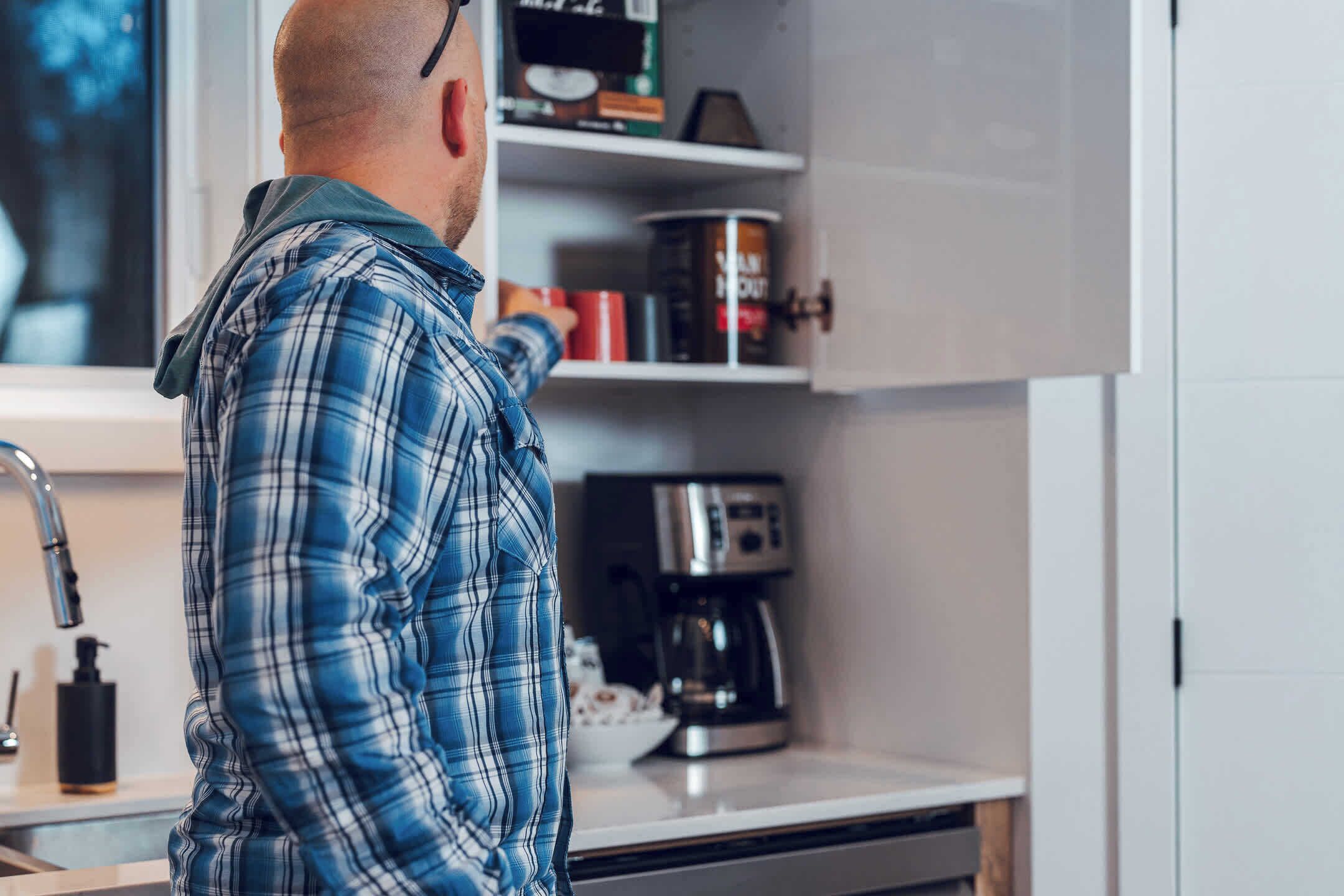 Una persona con camisa escocesa parada en una cocina moderna con una cafetera y un estante con diversos objetos.