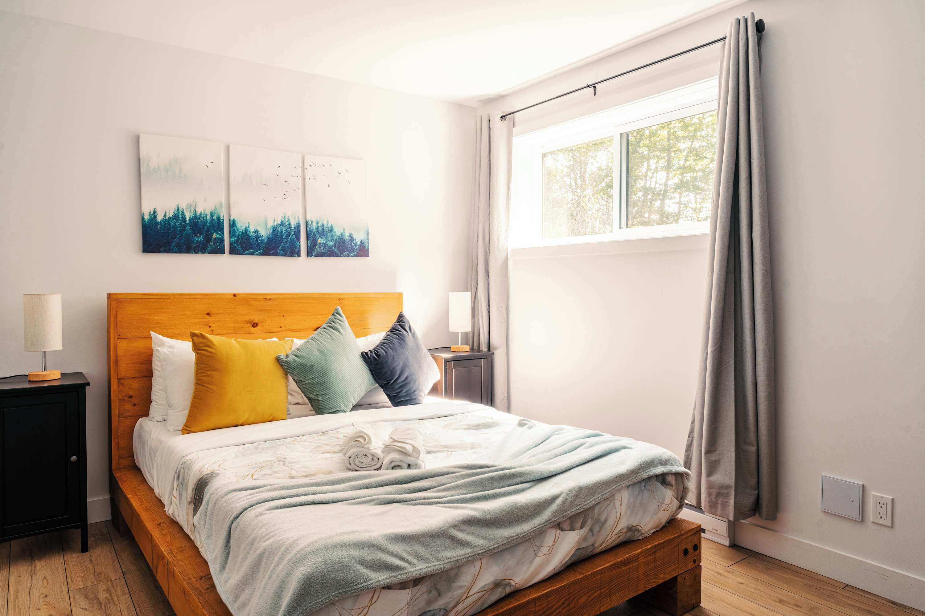A bedroom with a wooden headboard, a bed with a white and patterned bedspread, two bedside tables with lamps, and a window with curtains.