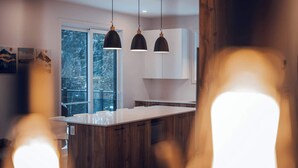 A modern kitchen with wooden cabinets, a large island, pendant lights, and a view of a snowy landscape through the window. - Yéti de Tremblant - Spa & Forest Charm (La Conception)