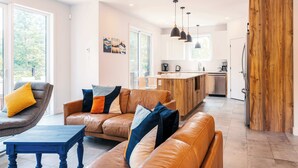A modern living room with leather sofas, a coffee table, pendant lights, and a kitchen area with wooden cabinets and a large window. - Yéti de Tremblant - Spa & Forest Charm (La Conception)