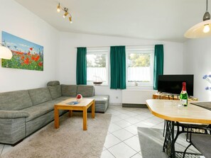 Uma sala de estar com um sofá cinza, uma mesa de centro de madeira, uma mesa de jantar com cadeiras, uma televisão e um quadro de flores.