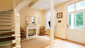 A staircase with a white balustrade leading to a landing. There is a white console table with a mirror above it, a wicker chair, and a door with a window to the right. - Hortensie by Interhome (Zempin)