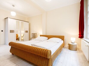 Une chambre à coucher avec un lit en osier, du linge de lit blanc, une petite table de chevet avec une lampe, une armoire et une fenêtre avec des rideaux rouges.