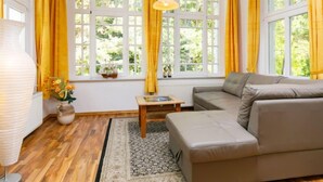 A cozy living room with a sofa, a small table, and a lamp. There are large windows with yellow curtains and a view of greenery outside. - Sonnenblume by Interhome (Zempin)