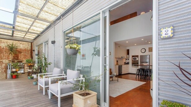 A modern patio with a glass door leading to a kitchen area, featuring a wooden deck, outdoor seating, and potted plants.