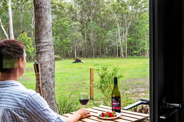 Una persona sentada a una mesa con una botella de vino y un plato de comida, con vistas a una zona verde con árboles al fondo.