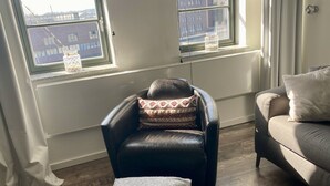 Ein Wohnzimmer mit Ledersessel, grauem Hocker und einer Couch. Zwei Fenster bieten Blick auf Häuser, ein kleiner Tisch mit Kerze steht daneben.