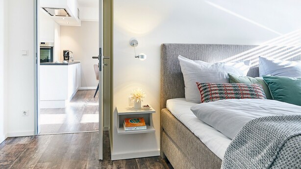 Ein modernes Schlafzimmer mit Bett, Nachttisch und Blick auf eine Küche.