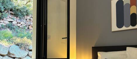 A hotel room with a bed, a nightstand, a lamp, a window with a view of greenery, and a wall-mounted artwork.