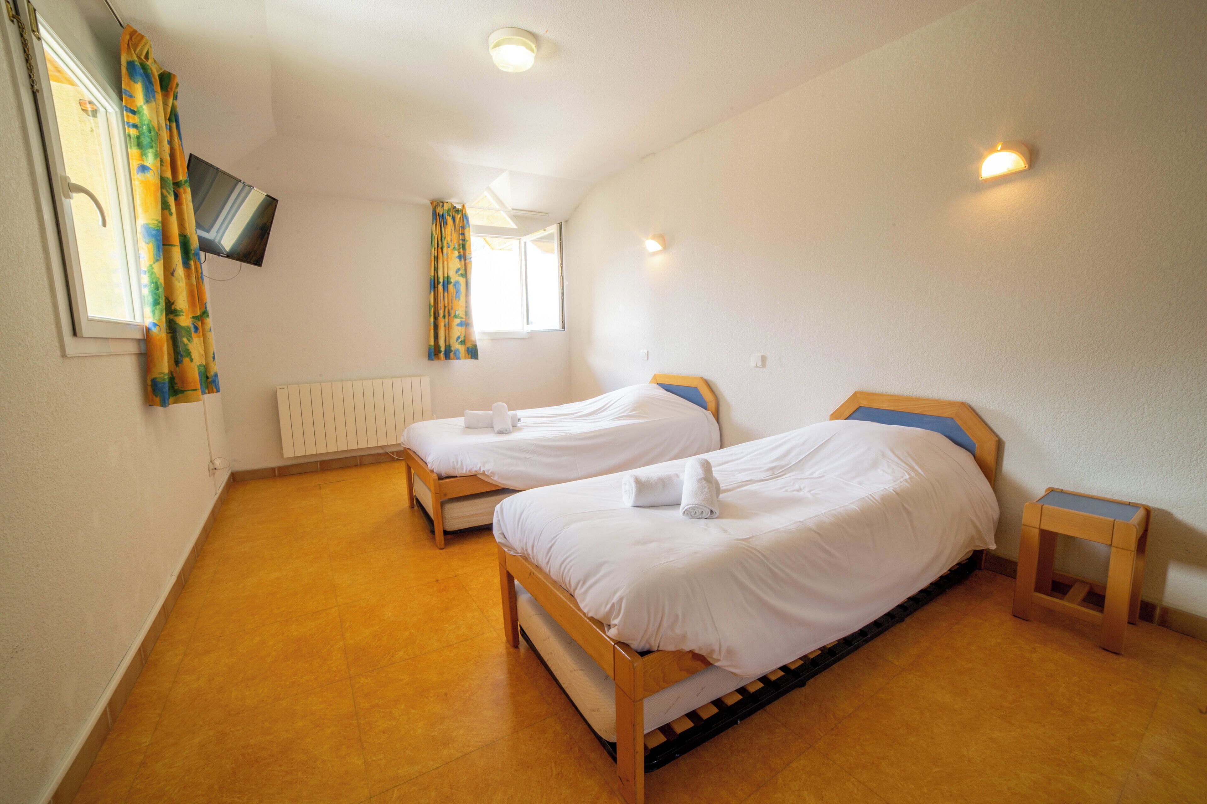 A room with two wooden beds, a small wooden table, a wall-mounted TV, and a window with curtains.