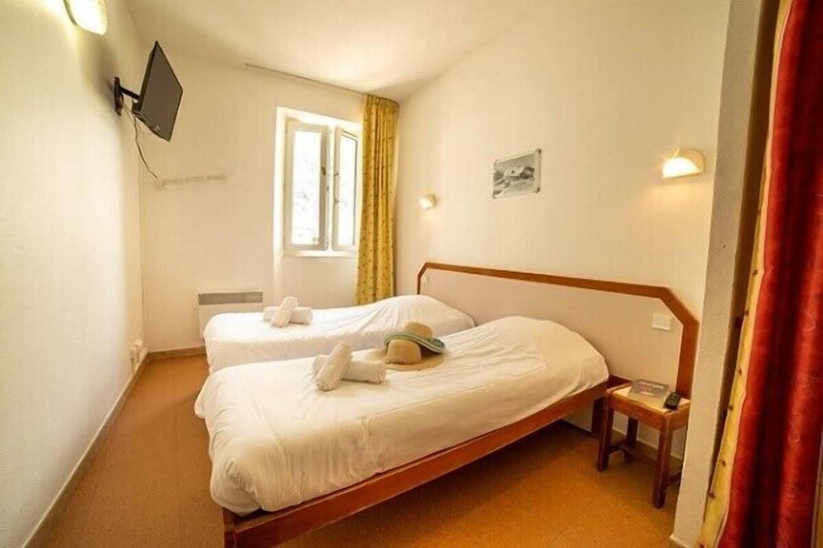A hotel room with two single beds, a small bedside table, a wall-mounted TV, a window with curtains, and a wall-mounted light.