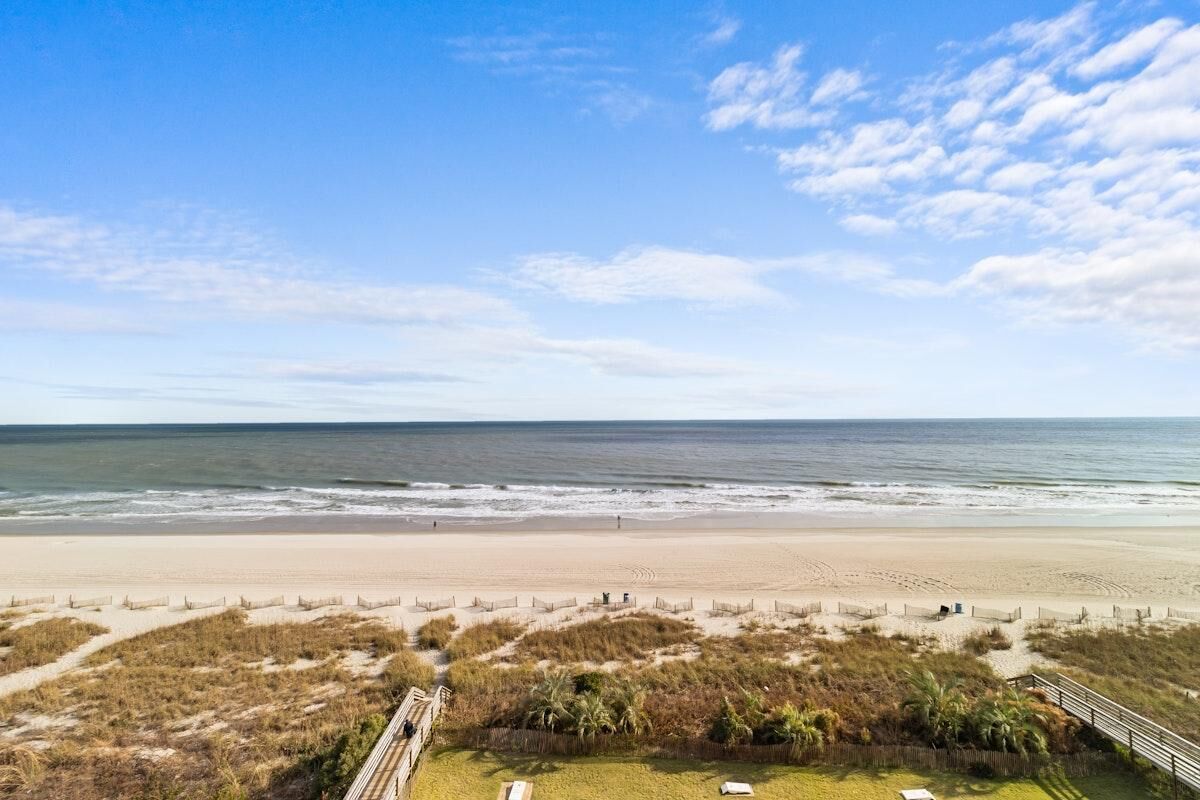 A beach view with a wooden walkway leading to a grassy area with a few chairs.
