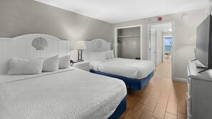 Quarto de hotel com duas camas, televisão e vista para o mar através de uma porta aberta.
