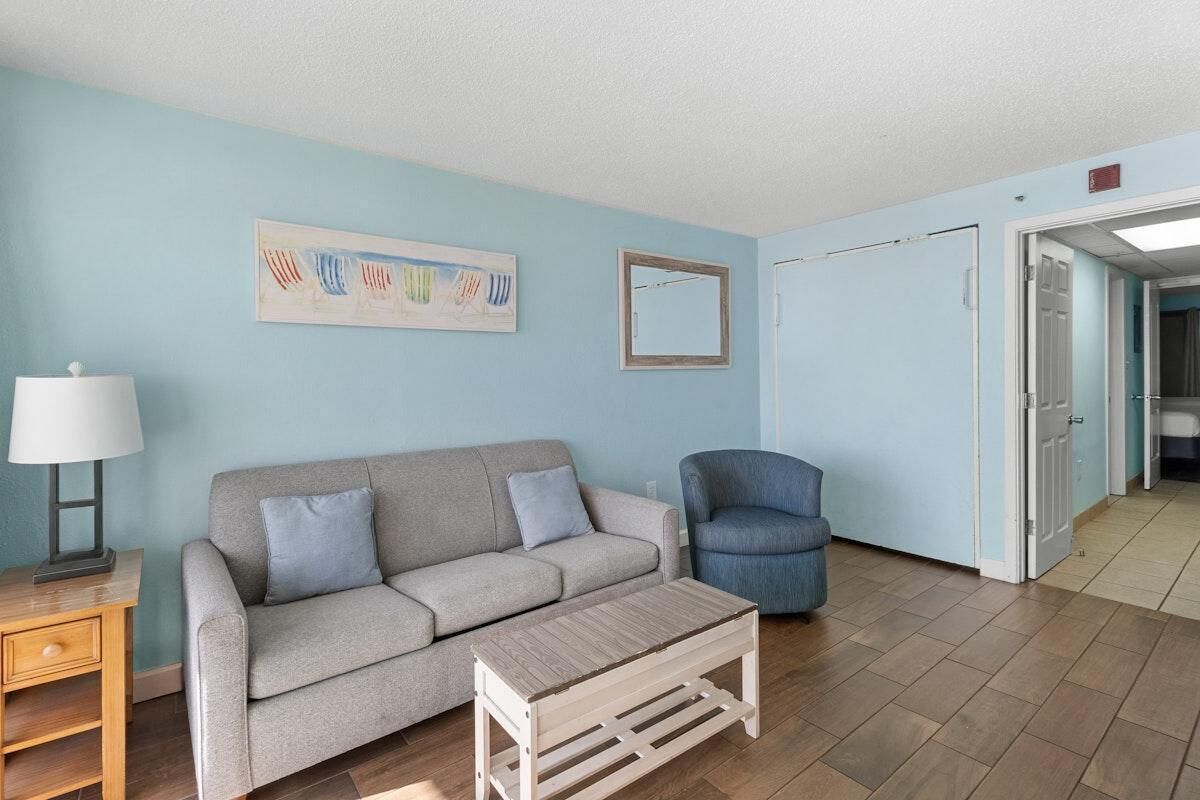 A living room with a grey sofa, a blue armchair, a wooden coffee table, a lamp, and a painting on the wall.