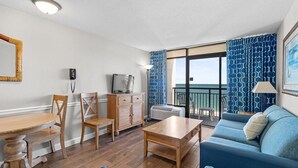 A living room with a wooden dining table, blue sofa, and a TV. - Beachy Oceanfront Suite with Breakfast 8th Floor (Myrtle Beach)