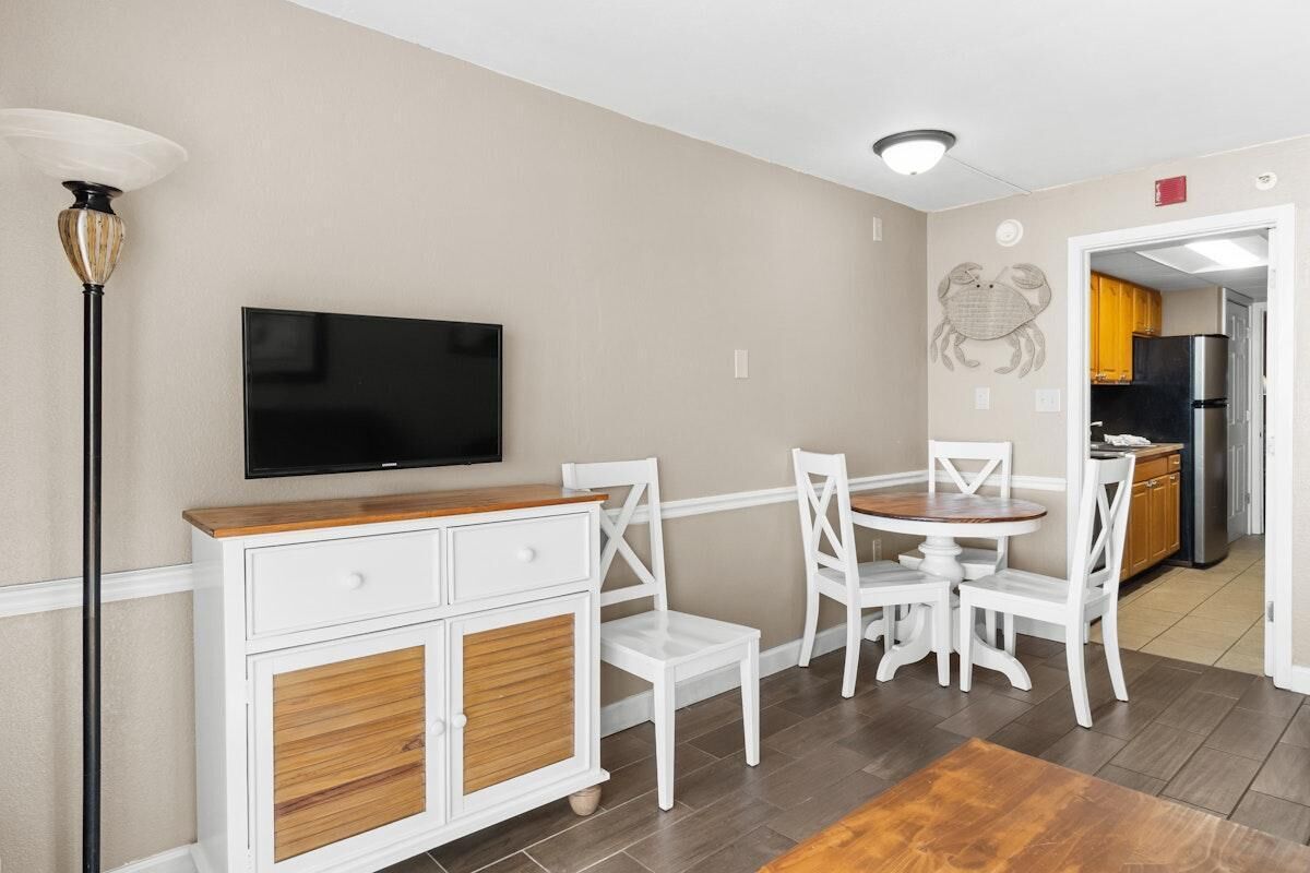 A dining area with a TV mounted on the wall, white chairs, a wooden table, and a cabinet with drawers.