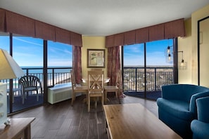 A room with a wooden floor, a blue sofa, a wooden table, and a chair. There is a window with a view of the ocean.