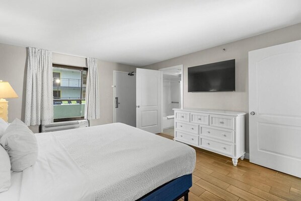 Ein Schlafzimmer mit Bett, Kommode, an der Wand befestigtem Fernseher und einem Fenster mit Vorhängen.
