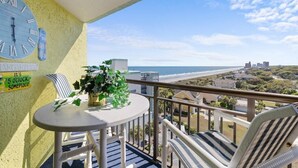 A balcony with a clock, a table with a plant, striped chairs, and a view of the beach and buildings. - Jaw-Dropping Ocean View King Ste w/Breakfast (Myrtle Beach)