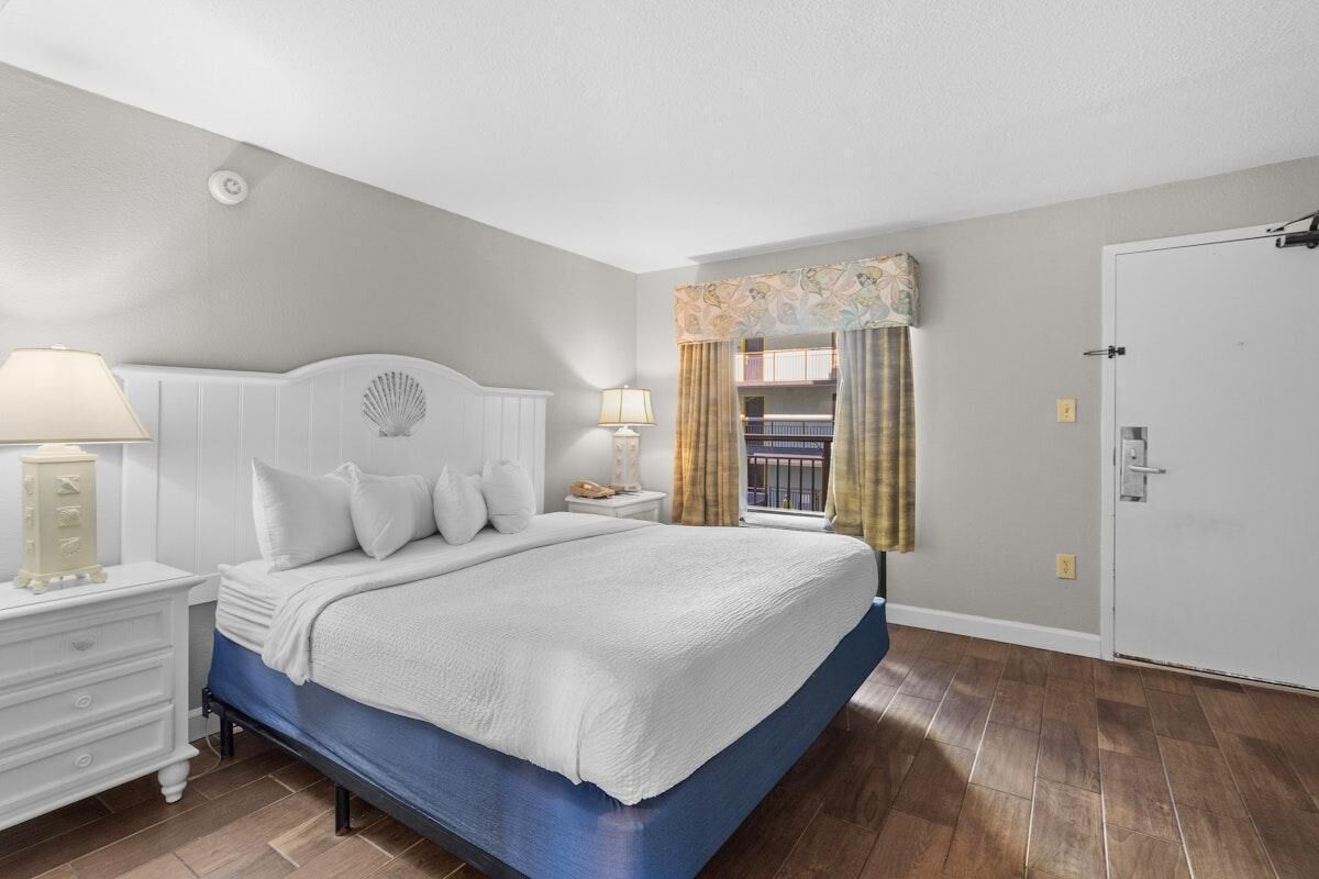 A bedroom with a large bed, two bedside tables, a nightstand, a window with curtains, and a door.