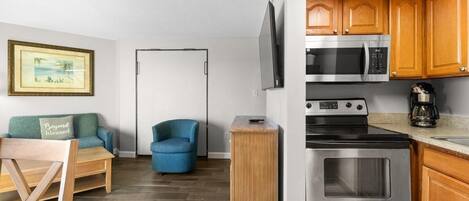 A compact kitchen with a microwave, oven, and coffee maker. There is a wooden cabinet and a blue chair.