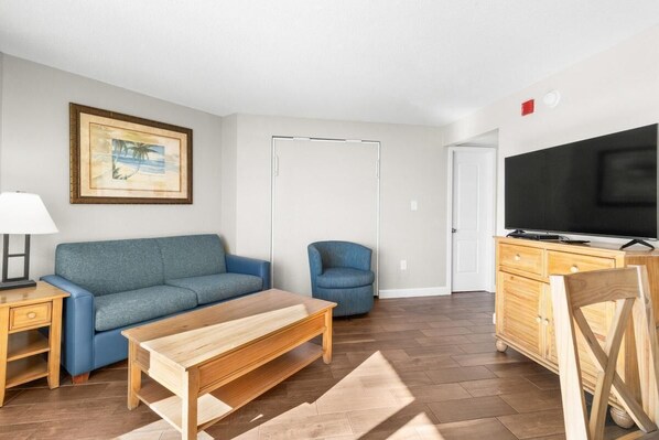 A living room with a sofa, a small chair, a wooden table, a TV on a wooden cabinet, and a framed picture on the wall.