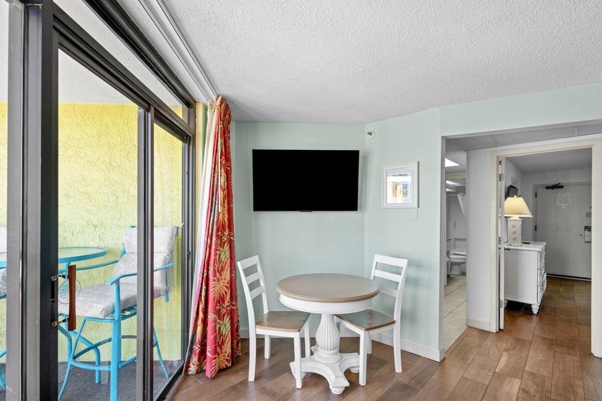 Une pièce dotée d’une porte-fenêtre coulissante donnant sur un espace extérieur, d’une table ronde avec des chaises, d’un téléviseur fixé au mur et d’une petite armoire avec une lampe.