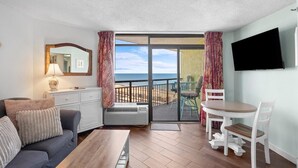 Ein Zimmer mit einer Couch, einem Tisch, einem Fernseher, einem Balkon mit Meerblick und einer Schiebetür.
