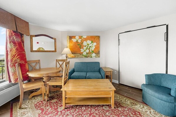 Ein Zimmer mit einem Holztisch, zwei Sofas, einem Esstisch mit Stühlen, einem roten Teppich mit Muster, einem Gemälde mit weißen Blumen, einem Spiegel und einem Fenster mit Blick.